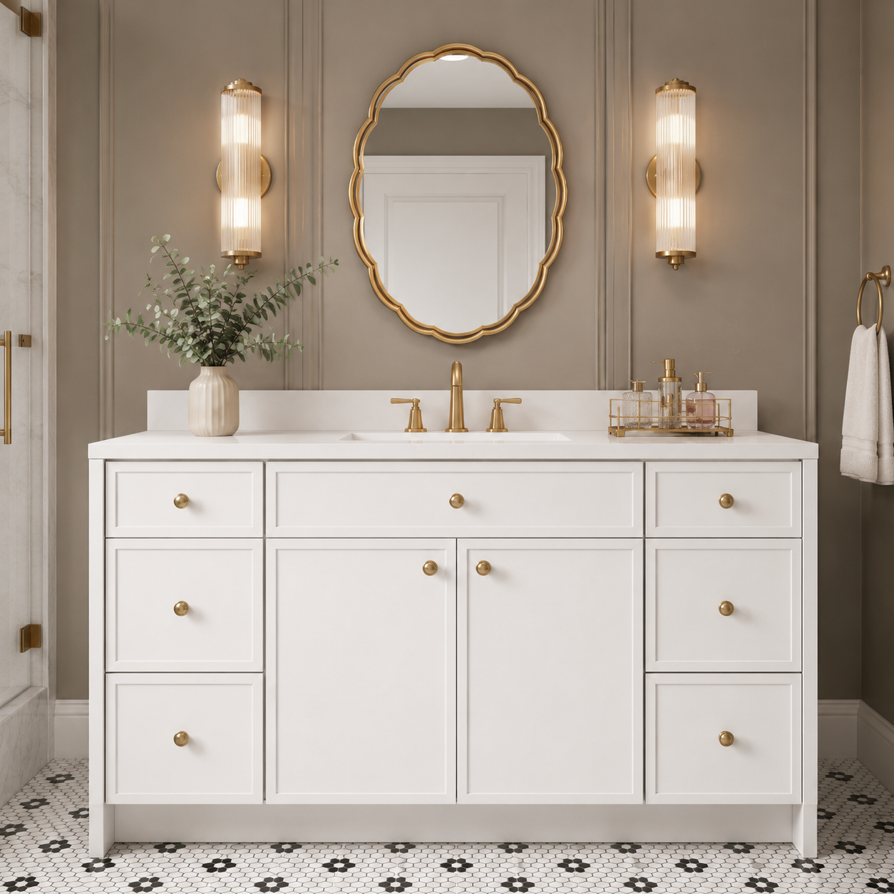 A white bathroom vanity with multiple drawers and cabinet doors, gold hardware, a white double sink countertop, a gold faucet, and a matching gold-framed oval mirror above. The vanity is styled in a bathroom with taupe paneled walls, gold sconces, a patterned tile floor, and a vase of greenery on the left.|USA Cabinets Outlet |